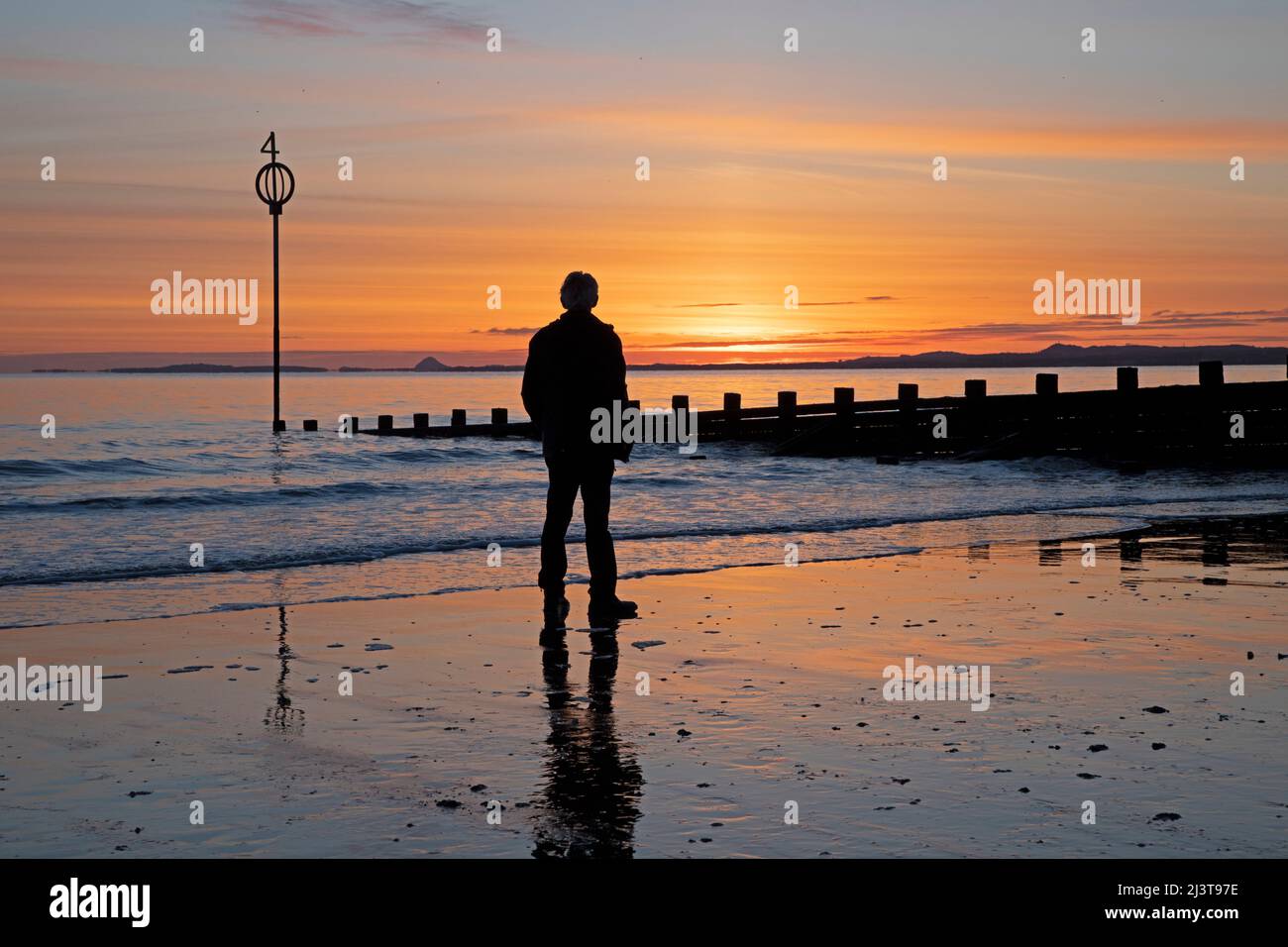 Portobello, Edinburgh, Scotland, UK. 10th April 2022.  Cool sunrise at minus 1 degrees centigrade beside the Firth of Forth. Pictured: a man stands on the shore looking towards the rising sun. Credit: Scottishcreative/alamy live news Stock Photo