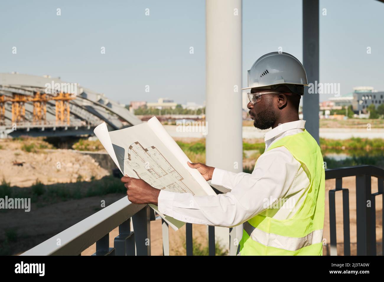 Engineer in hardhat and green vest viewing blueprint and standing on construction site Stock Photo