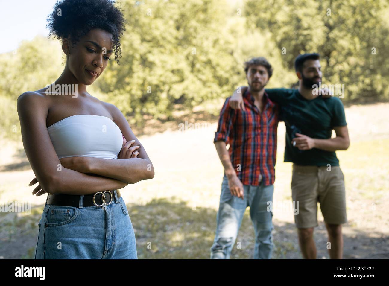 A girl of African descent with folded arms is bullied by two young men in an outdoor park. Concept of bullying, racism among young people. Stock Photo