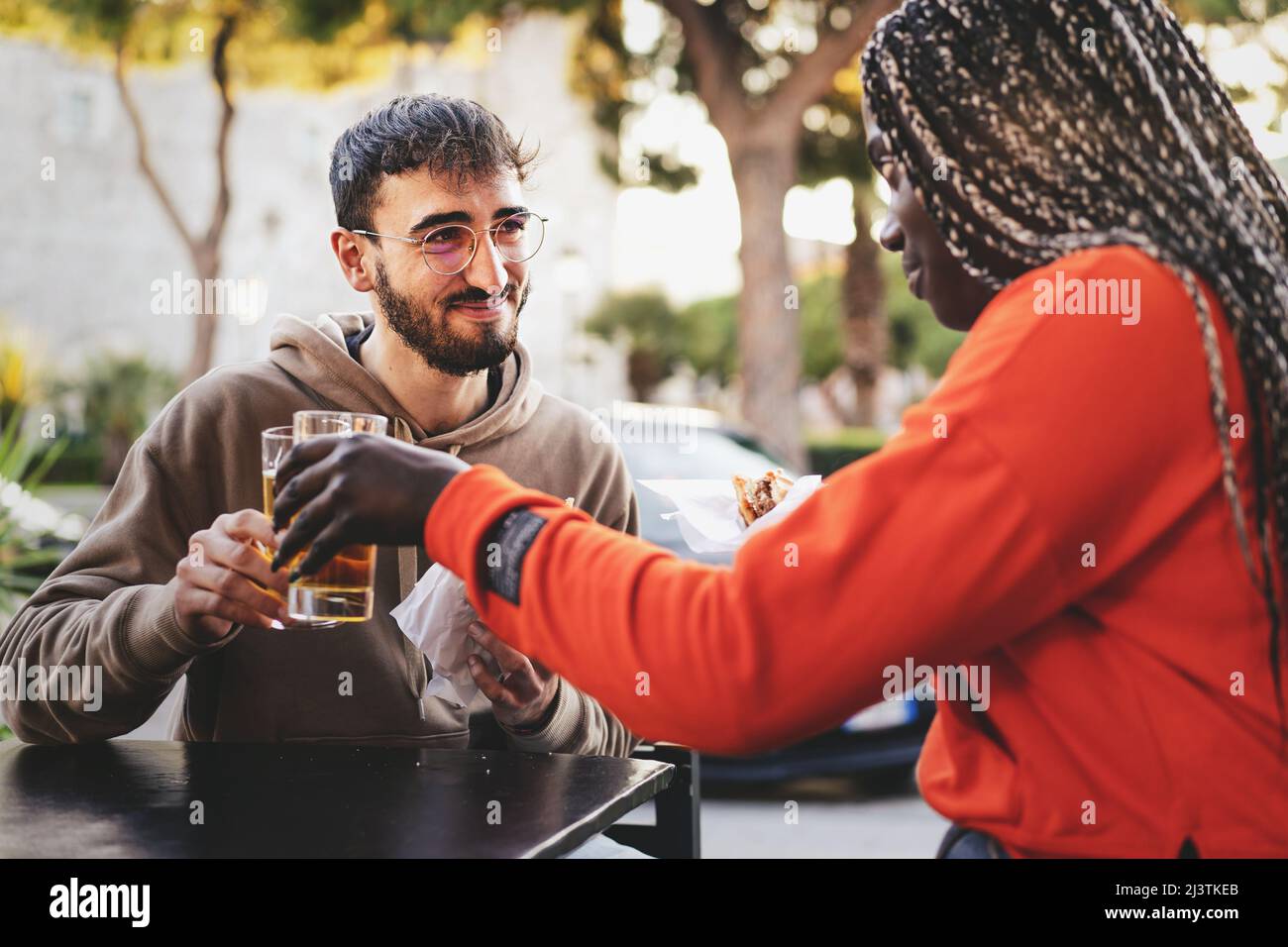 Biracial couple of young people sitting at cafe having a break - multicultural young couple toast with beers and eat some sandwiches Stock Photo