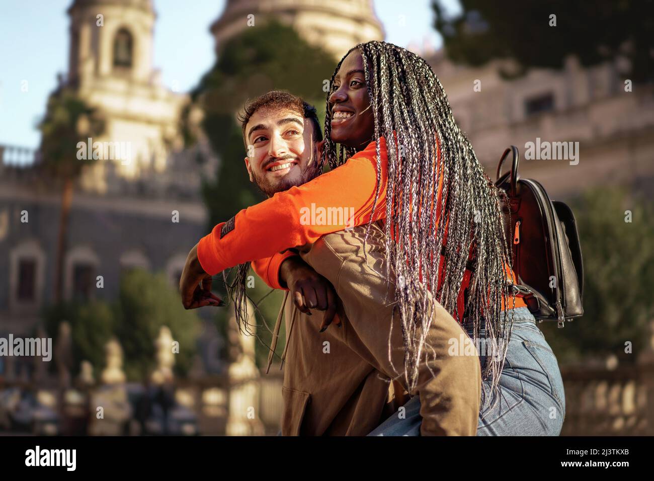 Cheerful couple of millenial travelers turning in the baroque city center piggybacking against a church background - International urban tourism in th Stock Photo