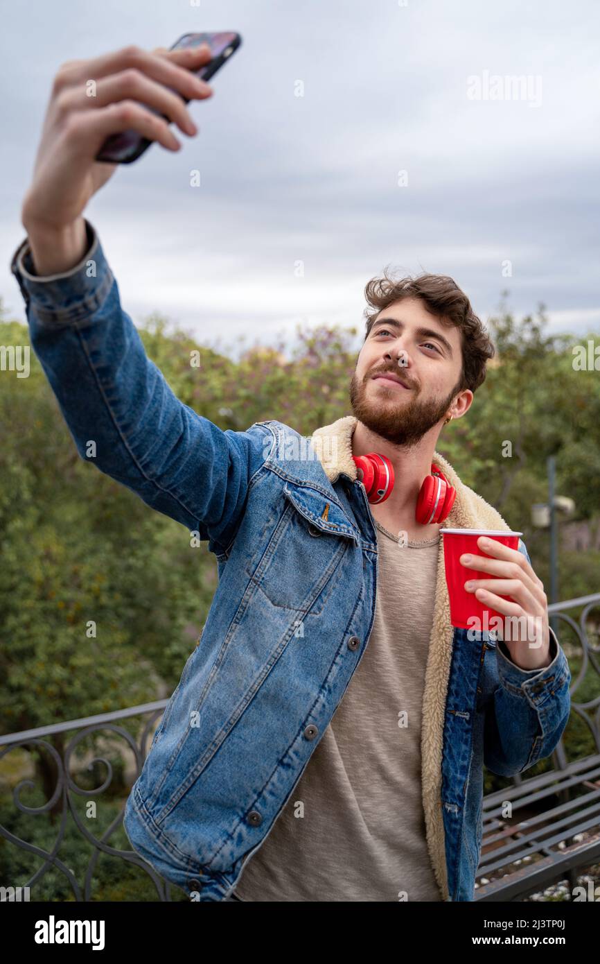 Young man with wireless headphone taking selfie outdoors. Gen z  people using tech concept. Stock Photo