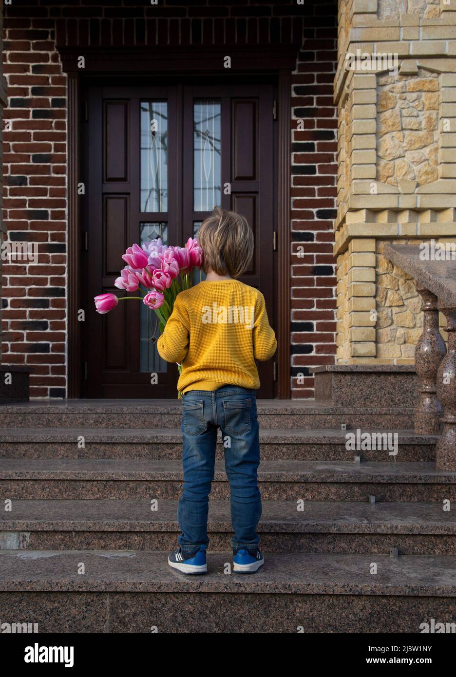 unrecognizable boy of 5-6 years old stands with his back with a bouquet of tulips in front of the door of the house. Festive mood, a gift with love fo Stock Photo