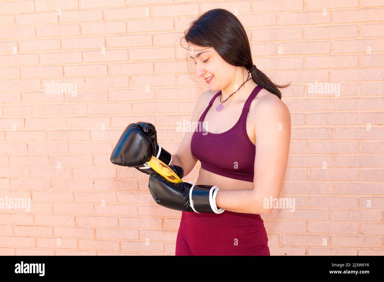 A young Caucasian woman is wearing black boxing gloves and trying to peel a banana. In the background is a brick wall. Stock Photo