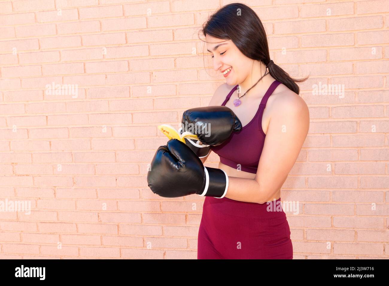 A young Caucasian woman is wearing black boxing gloves and has managed to peel a banana. In the background is a brick wall. Stock Photo