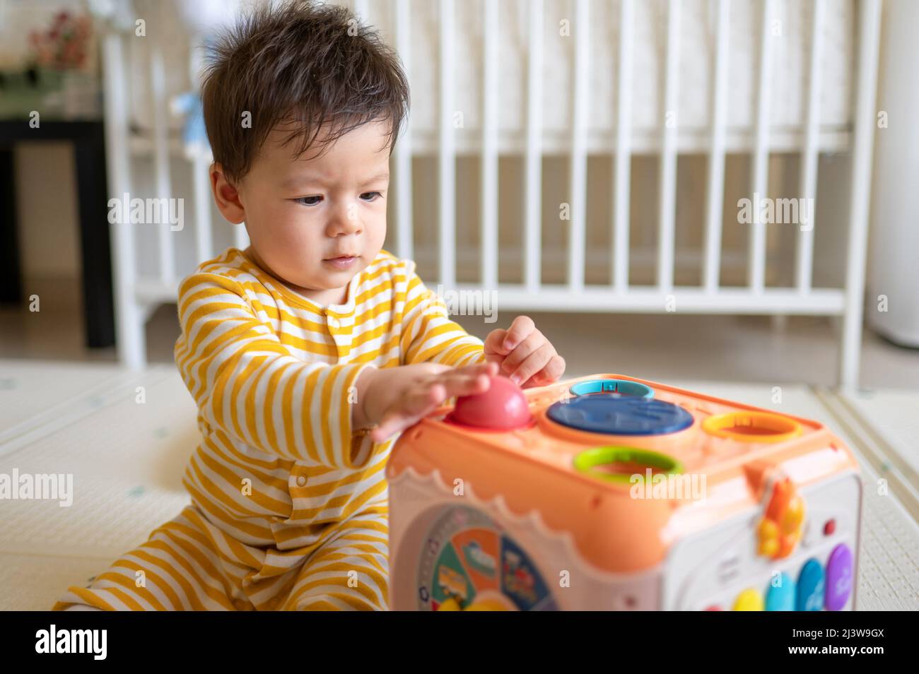 8 months old mixed race baby boy playing with activity box in the bedroom wearing yellow pajama while sitting on the floor covered with a rubber play Stock Photo