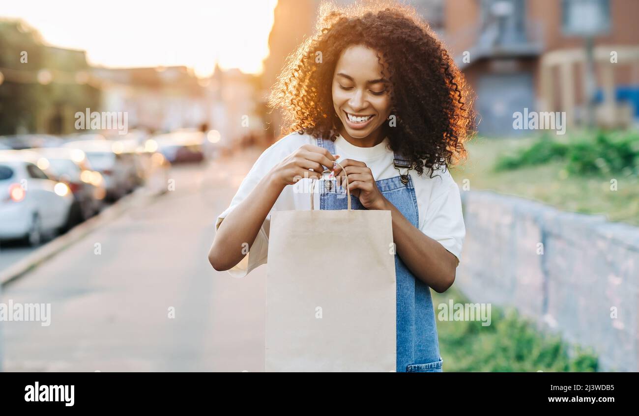 Takeaway and delivery food concept. Young african woman holds craft paper bag outdoors and look inside.  Stock Photo