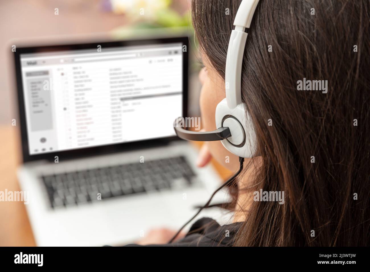 Home office, customer support, help desk concept. Woman with headset working with a laptop, talking on phone, view from behind. Stock Photo