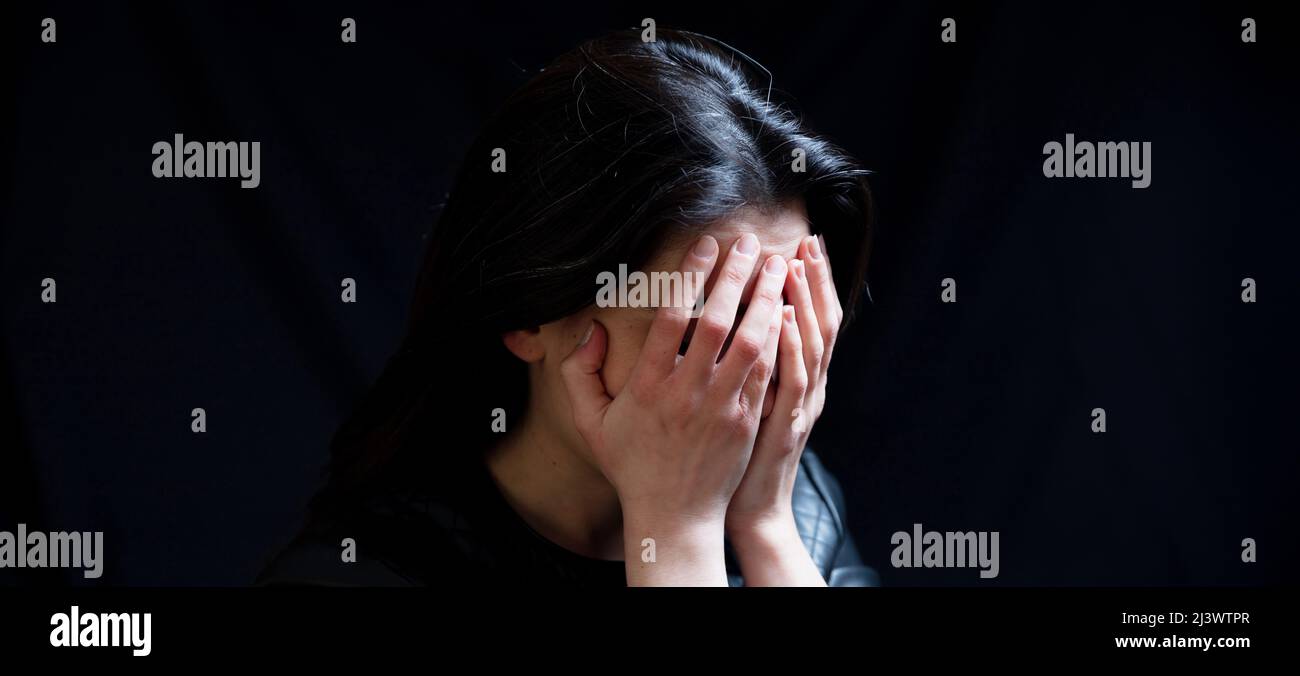 Young woman covering eyes, sad and frightened in a dark room, copy space. Fear and despair concept Stock Photo