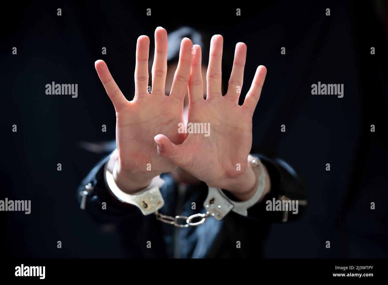 Young woman with raised handcuffed hands for protection. Dark room background, copy space. Hostage, captive and violence concept Stock Photo