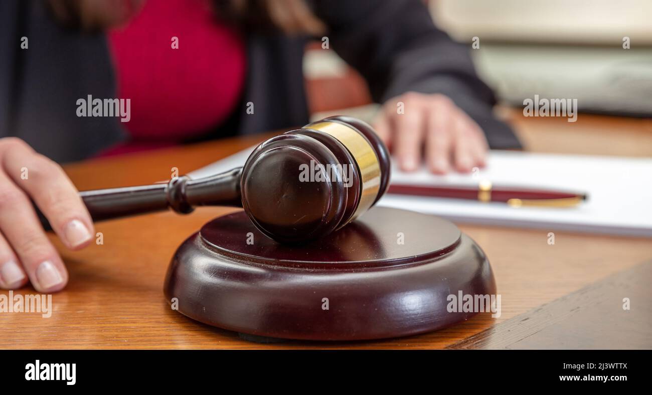 Judge gavel on a wooden table close up view. Woman sitting with crossed hands and a notepad infront her, auction Stock Photo