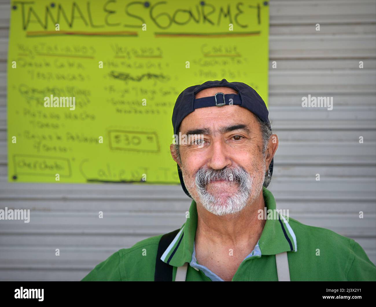 Positive Mexican man (cook, chef) sells traditional food (tamale) at the slow food market, stands in front of hand written menu and looks at viewer. Stock Photo