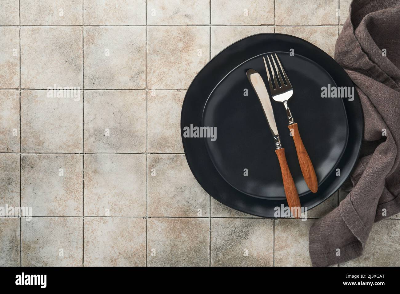 Ceramic empty black plate, silverware and linen kitchen towel napkin on old ceramic tile table background. Cooking stone backdrop. Top view with copy Stock Photo