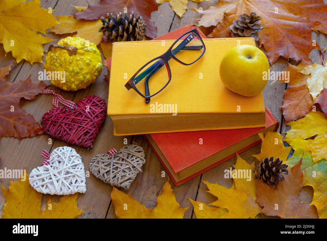 Autumn background. Old books, apple, glasses and decorative hearts on wooden planks with autumn leaves. Autumn design concept. Stock Photo