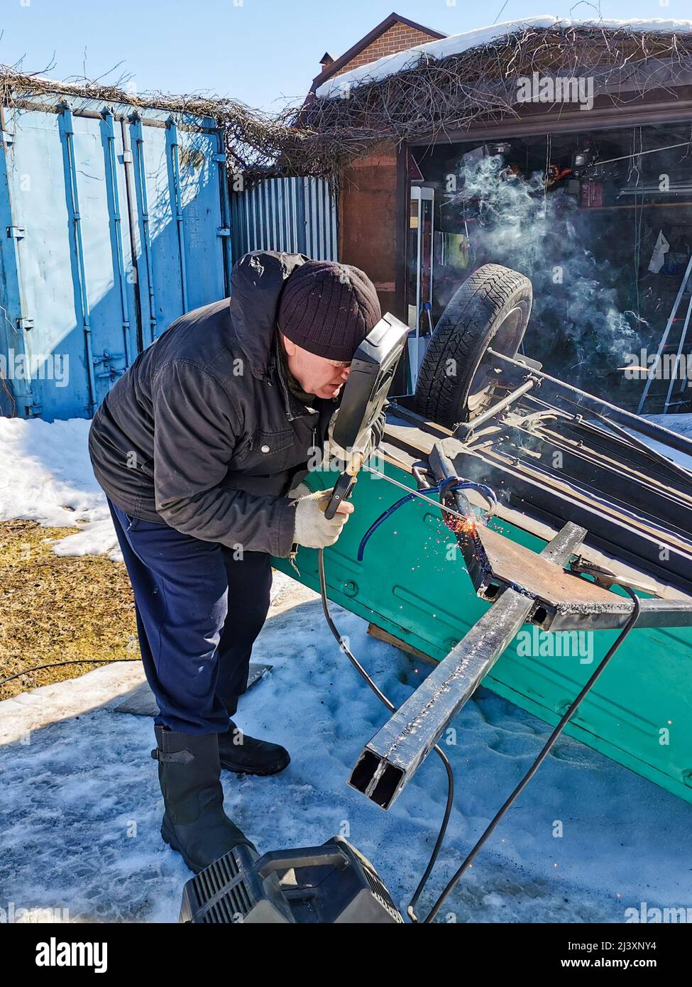 An elderly male welder repairs a car trailer with electric arc welding