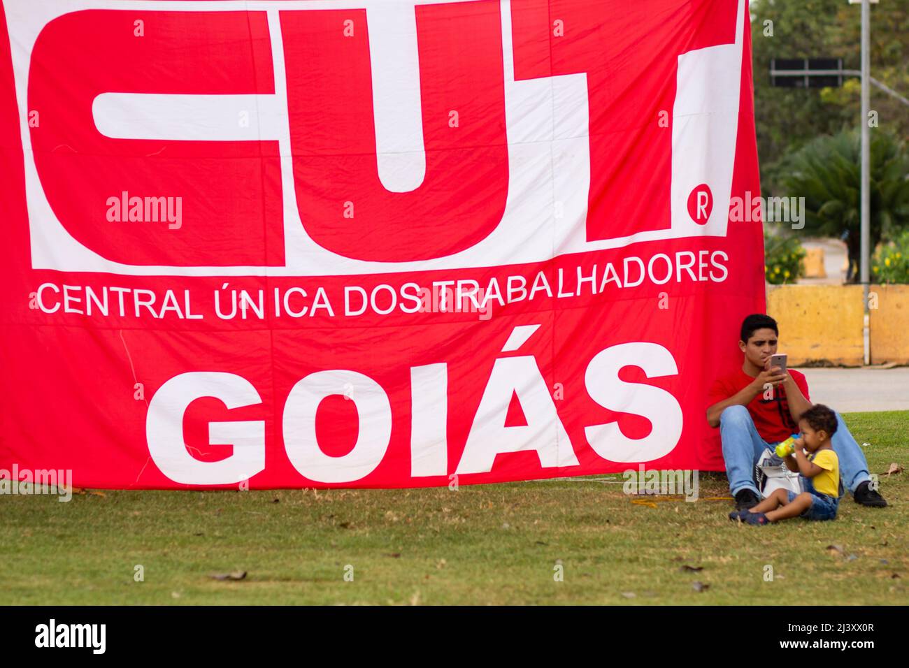Goiânia, Goias, Brazil – April 09, 2022:  A man with a child sitting under a CUT flag. Photo taken during a protest, in the city of Goiânia. Stock Photo