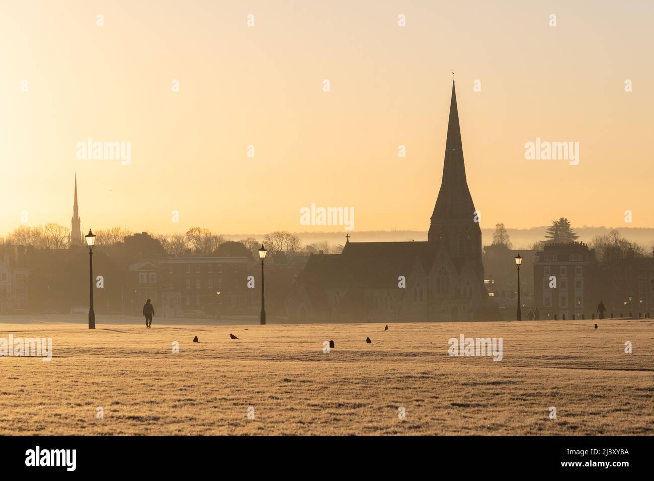 A view of Blackheath Common in the winter during a warm sunset. All Saints church can be seen in the distance and people can be seen walking. Stock Photo
