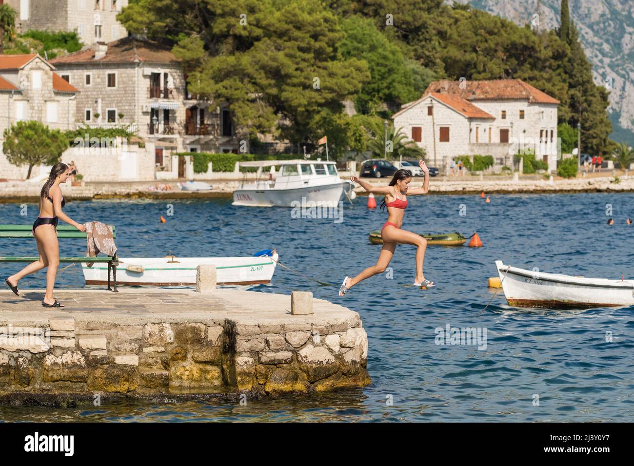 Two girls jumping from a stone pier in Perast village in Kotor bay ...