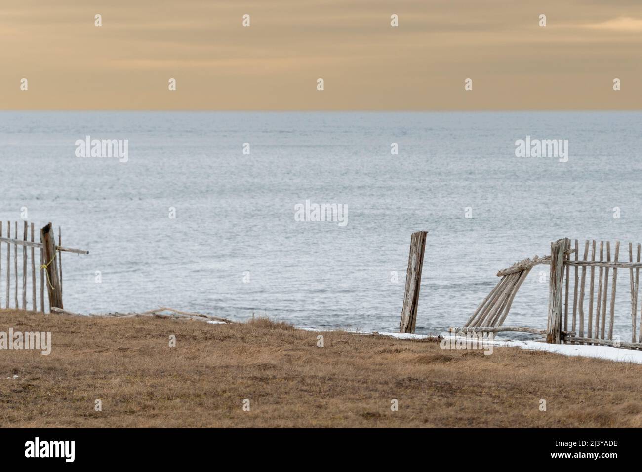 A broken fence on the edge of a cliff overlooking the ocean. The wooden fence is made of wood sticks. The ocean is calm and pale blue and sky yellow. Stock Photo