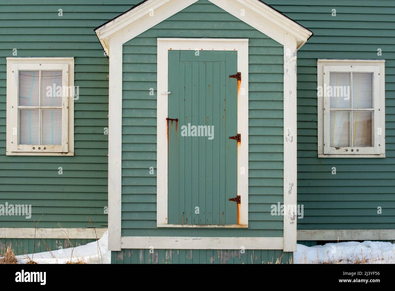 The exterior facade of a vintage wooden building. There's a green single shutter door with rusty hinges, white trim and wood boards on the house.. Stock Photo