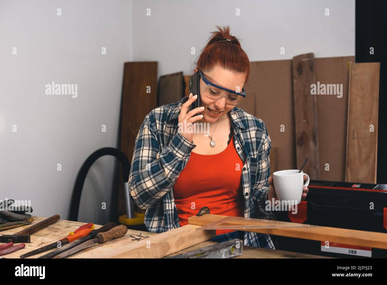 Young red-haired female carpenter talking to a customer on her mobile phone, having a cup of coffee in the morning. Stock Photo