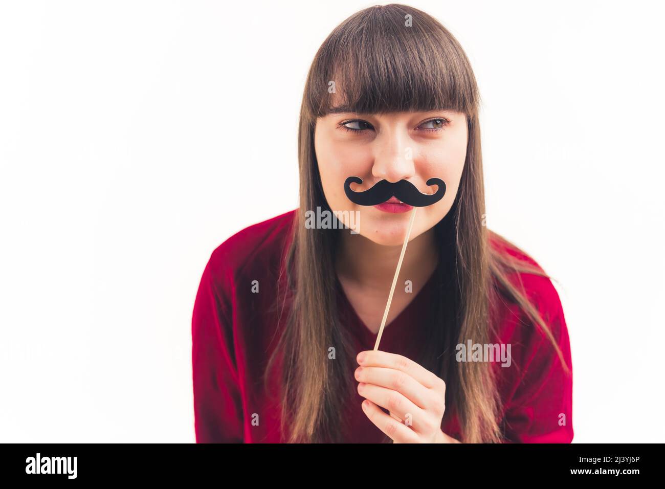 Pretty young woman holds fake moustache in front of her face and looks away movember concept white background copy space isolated studio shot . High quality photo Stock Photo