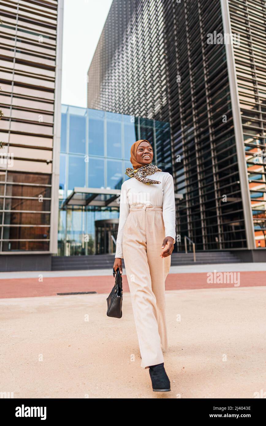 Full body of happy African American female wearing smart casual outfit and traditional headscarf walking with bag in central district against high tower and looking away with wide smile Stock Photo