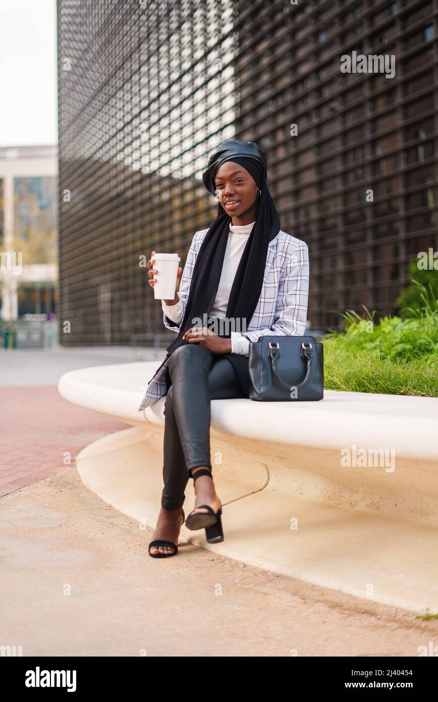 Full body of confident African American female in smart casual clothes and Muslim hijab sitting with crossed legs and having takeaway coffee in city park Stock Photo