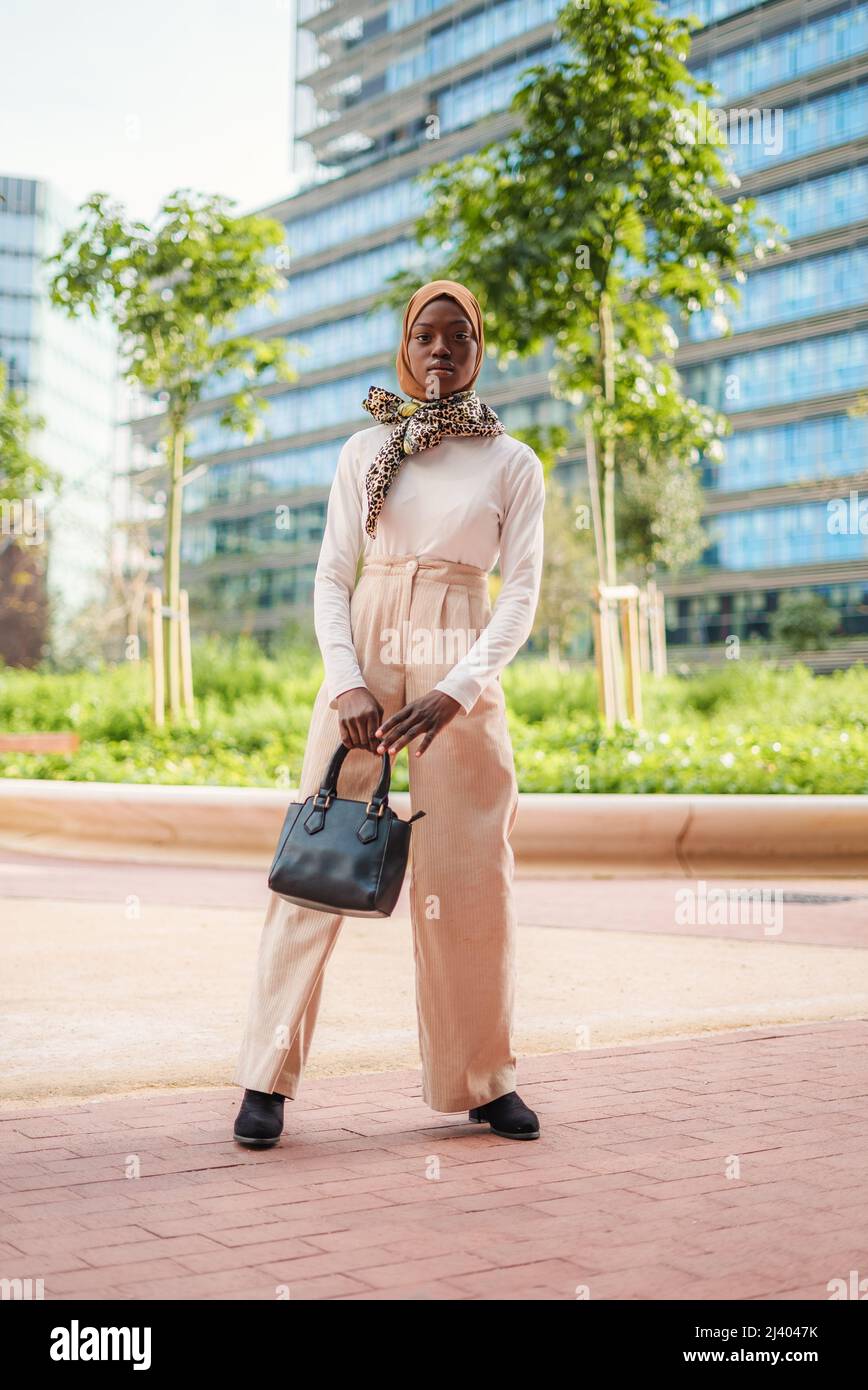 Full body of self assured female dressed in smart casual outfit and traditional Islamic hijab with scarf on neck standing in park and looking away Stock Photo