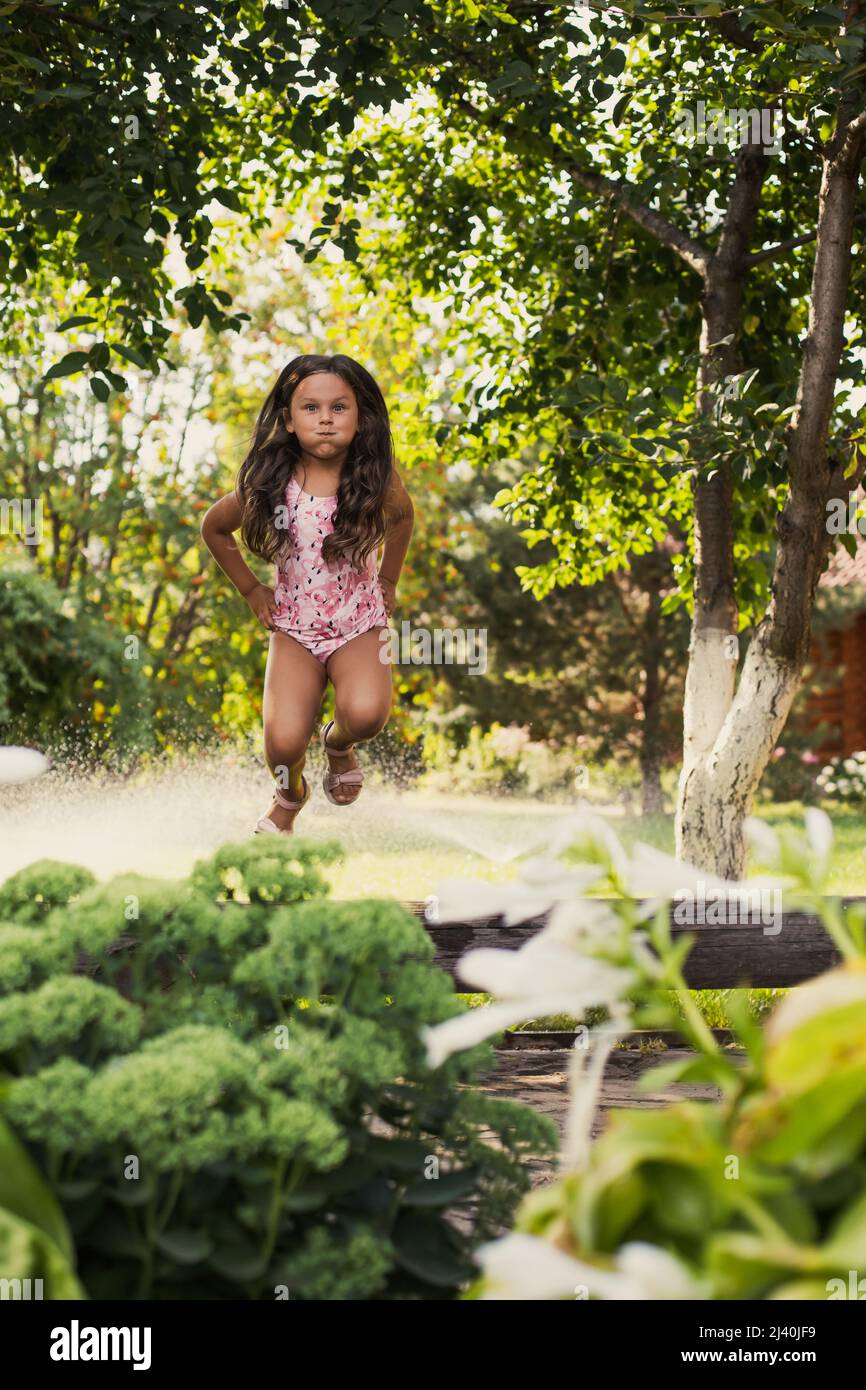 Playful blissful female child jumping off log with puffy cheeks, playing outside in park indulging with trees and bushes in background in daytime Stock Photo