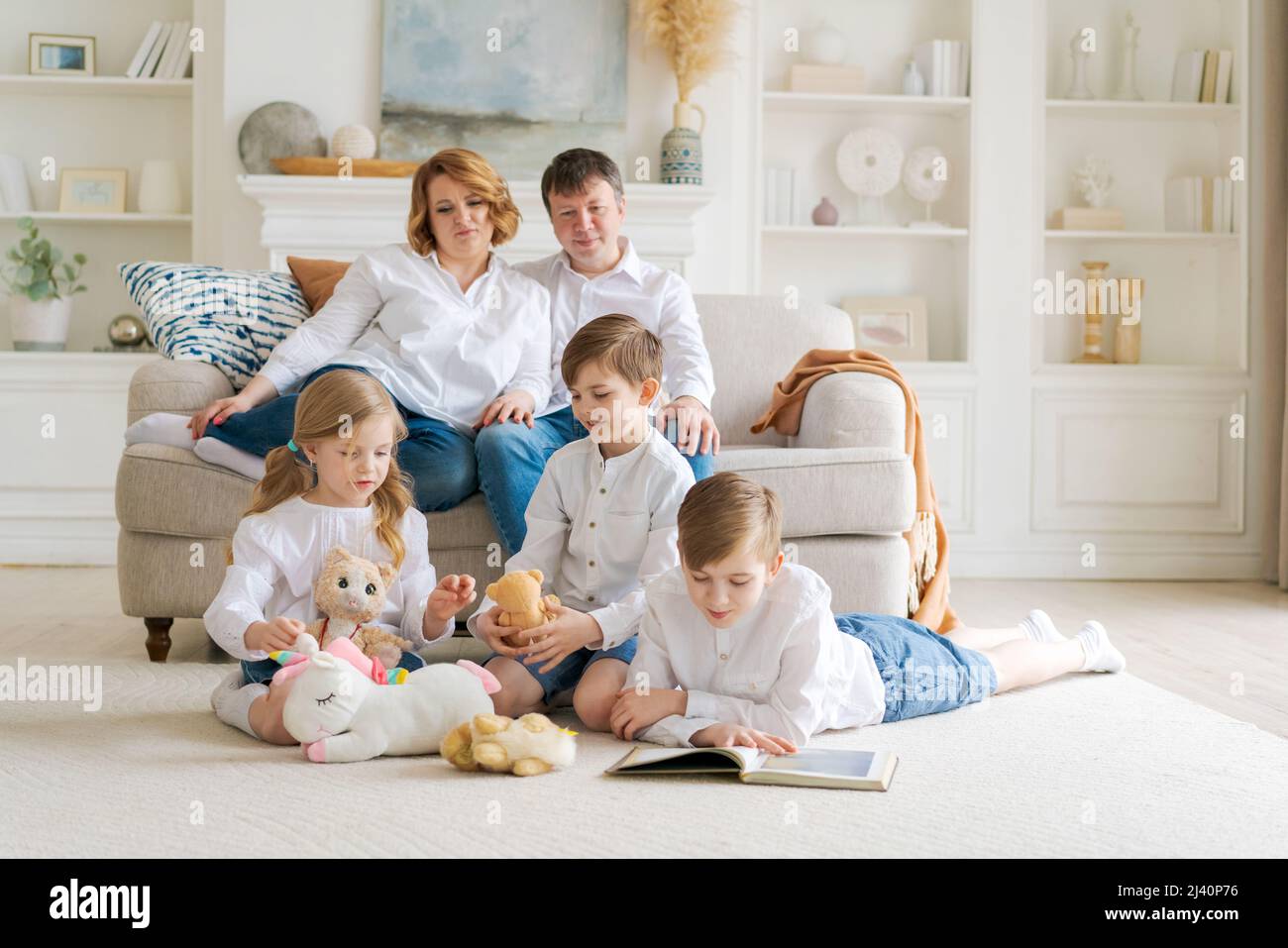 Happy young caucasian family resting in a new cozy design home on weekends. Delighted parents resting in their own home, children have fun sitting on the floor with toys and a book Stock Photo