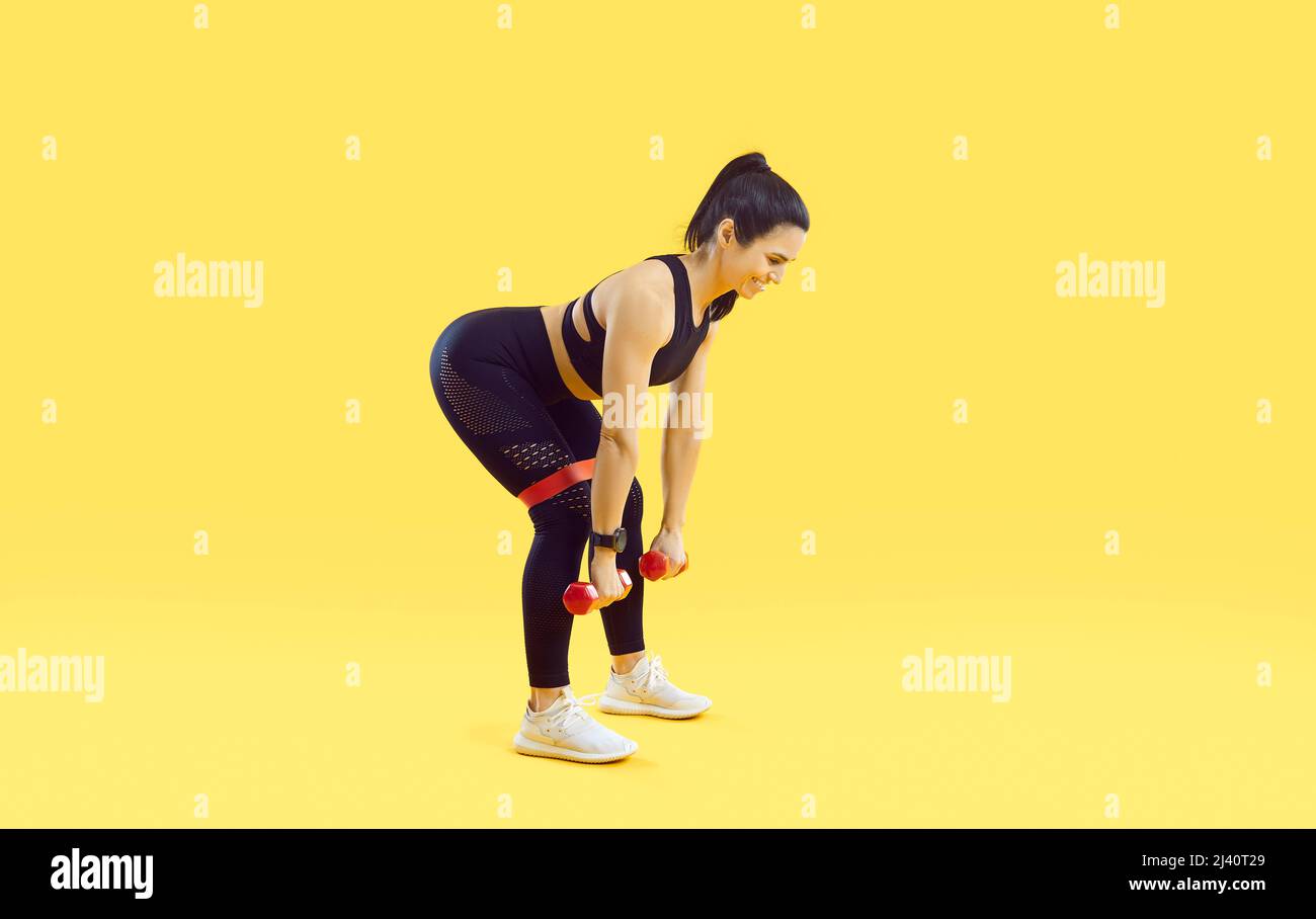 Woman doing sports exercises with dumbbells and with elastic fitness band on yellow background. Stock Photo