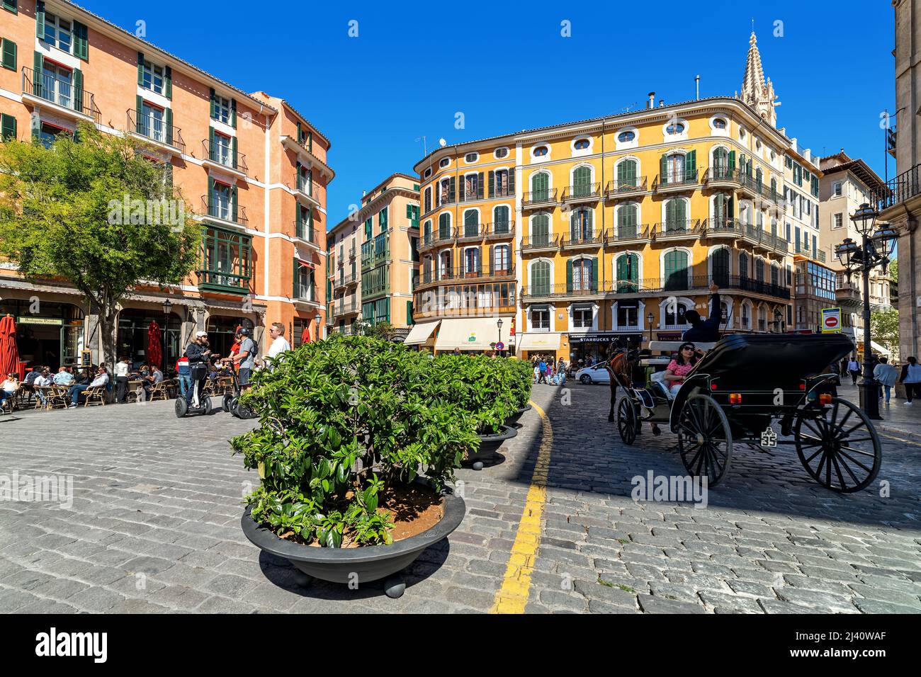 People and horse-drawn carriage on cobblestone street among colorful buildings in historic part of Palma, Spain. Stock Photo