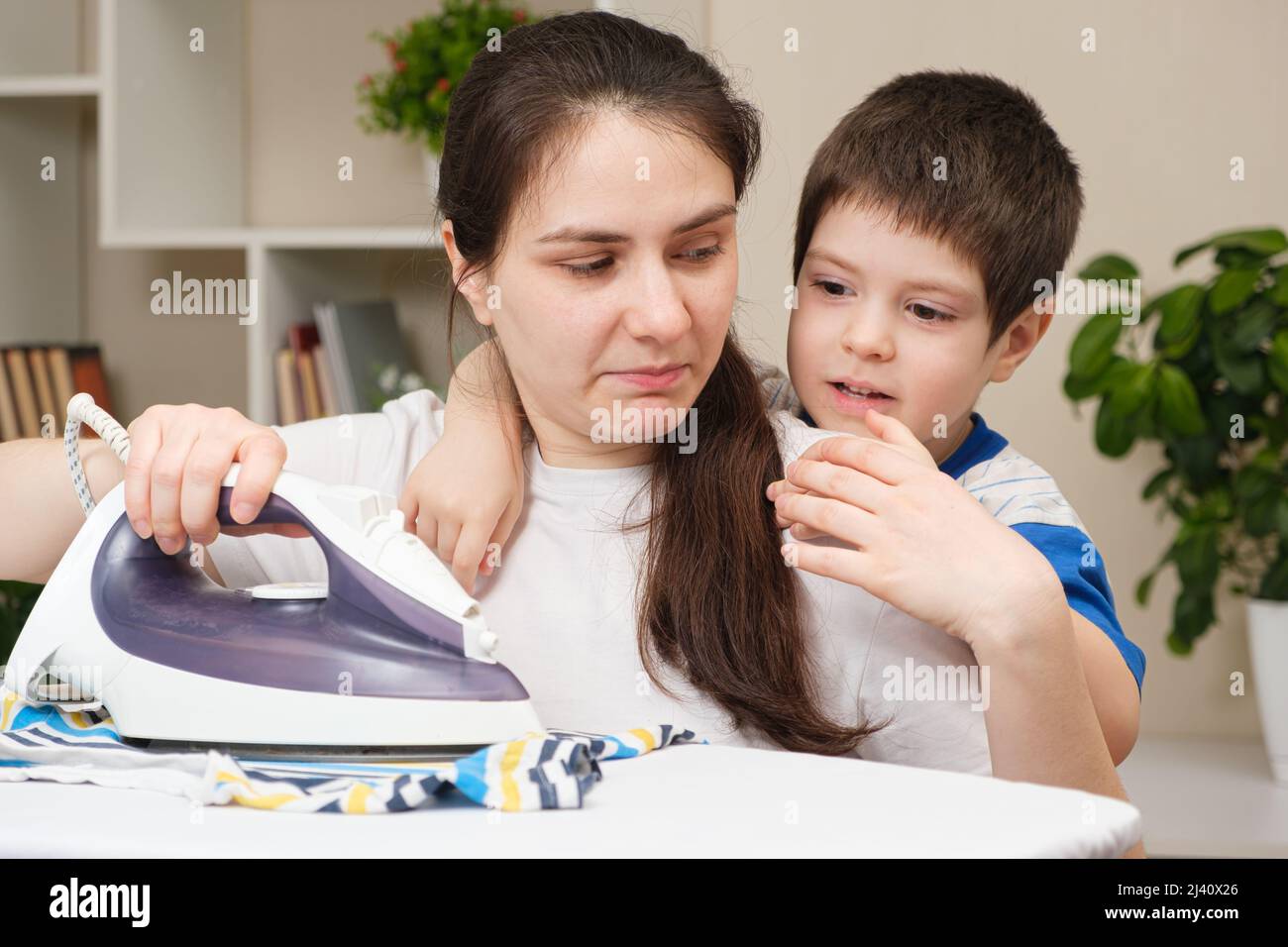 A small child of 4 years prevents his mother from ironing clothes. The difficulties of motherhood. Stock Photo