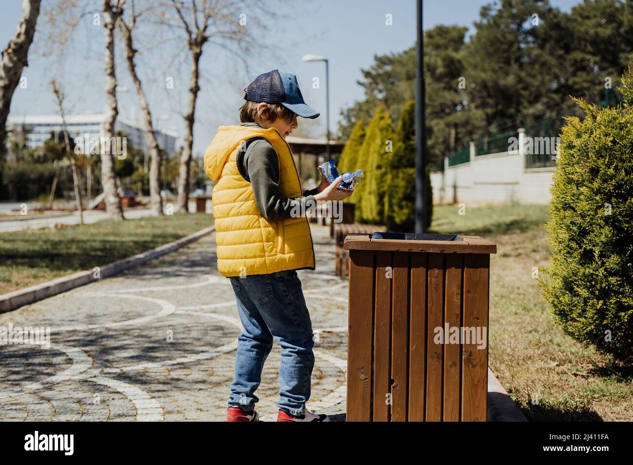 Schoolboy kid throwing the trash into dumpster. Boy using recycling bin to throw away the litter. Caucasian child recycles the junk into the trash-can Stock Photo