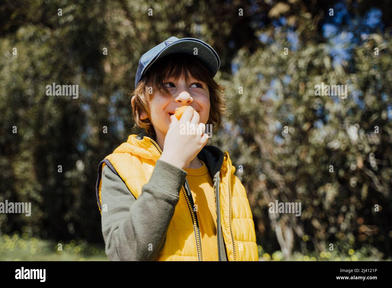 Smiling cheerful child kid in yellow vest and green hoodie eats crisp snacks outdoors in public park. Schoolboy boy enjoying consumes chews junk food Stock Photo