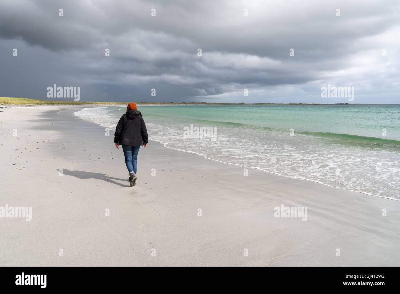 A woman walks along a deserted white sand beach backed by machair, with storm clouds on the horizon. Stock Photo