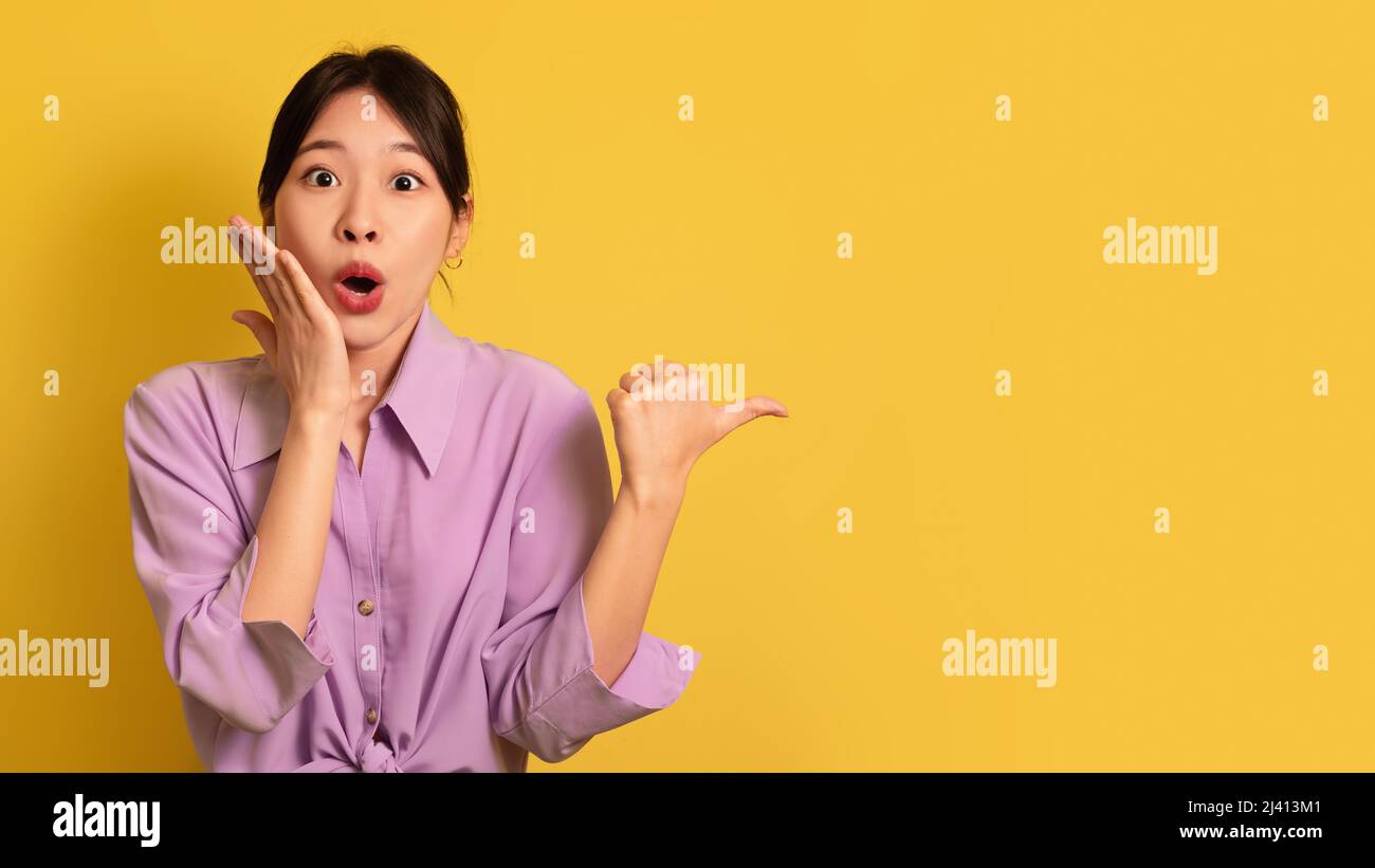 Overjoyed young Asian woman pointing aside with thumb up, shouting in ...