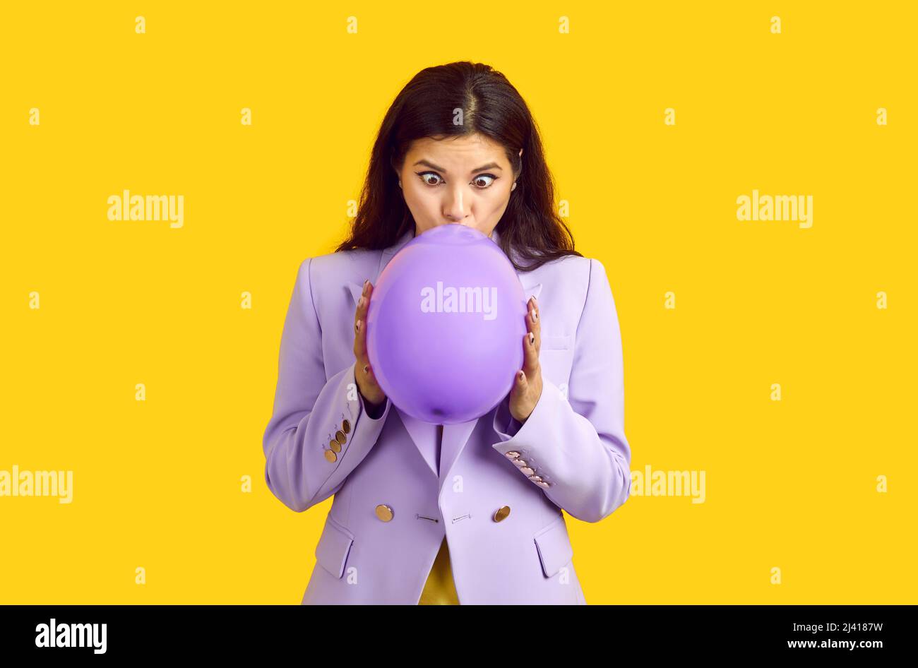 Beautiful woman isolated on yellow background inflating purple balloon with funny face expression Stock Photo