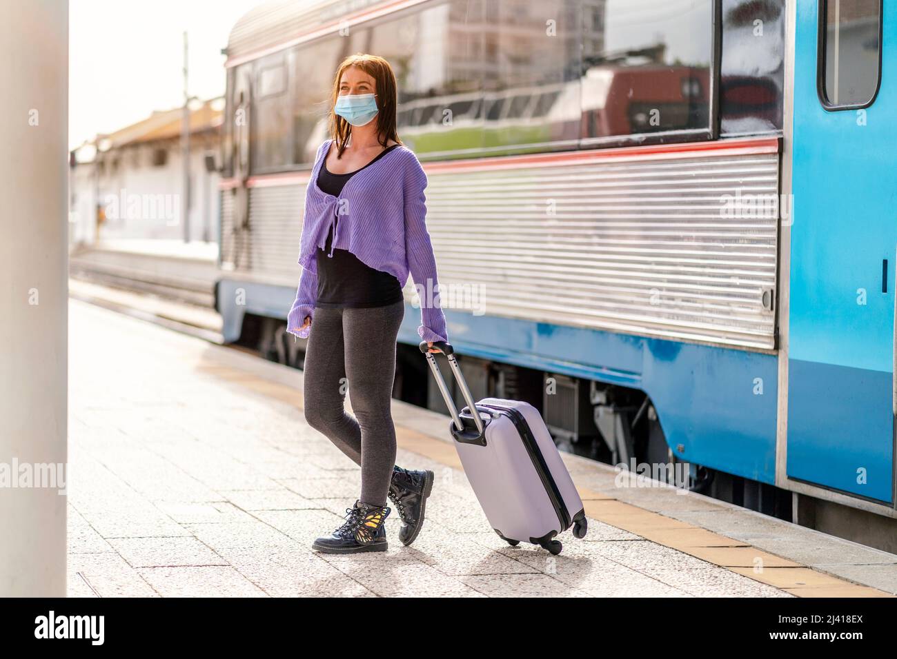 A young woman in a mask and with luggage walking on the platform of a train station at her destination Stock Photo