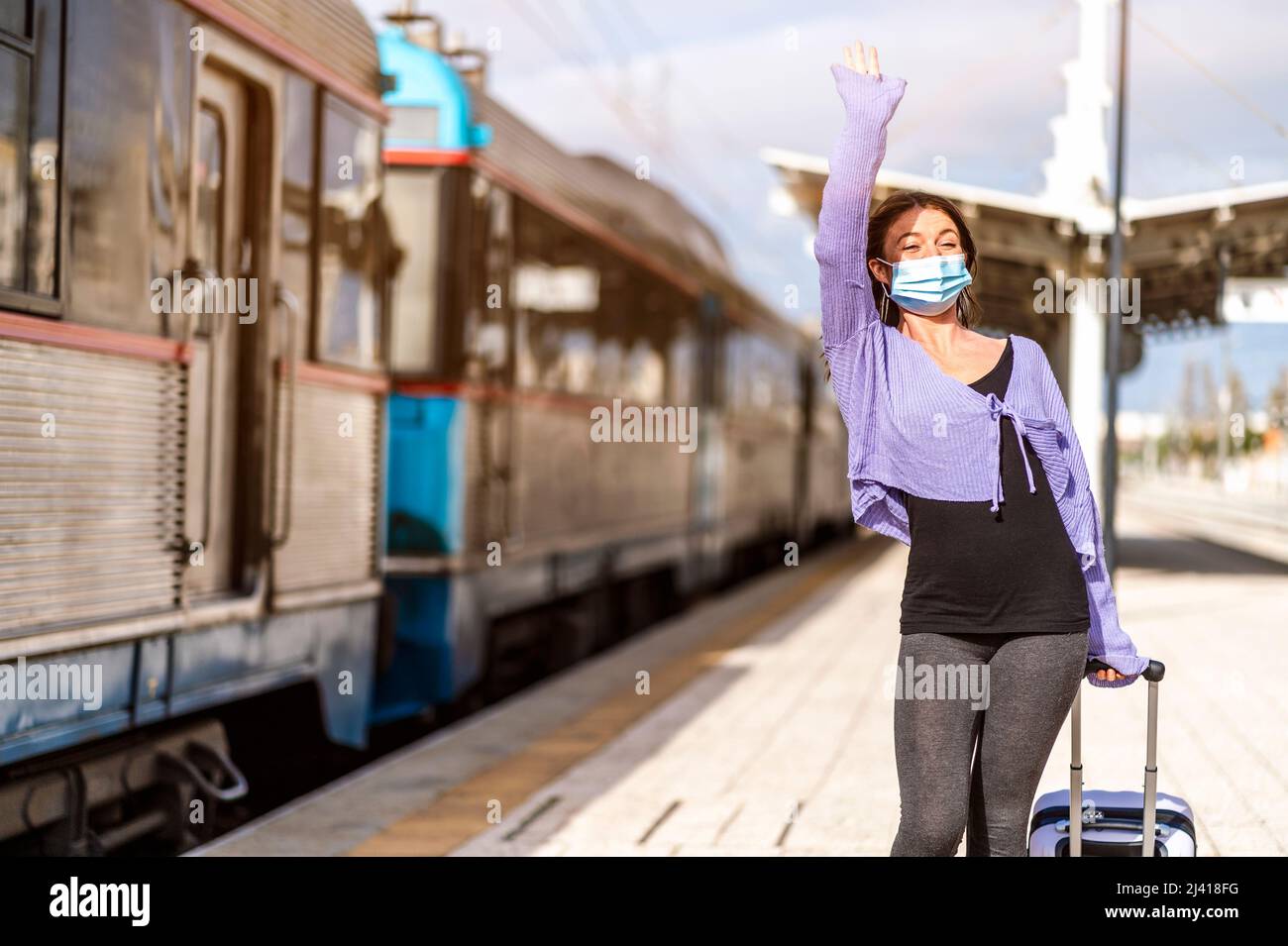 A young woman in mask and with luggage greetings somebody at the platform of train station Stock Photo