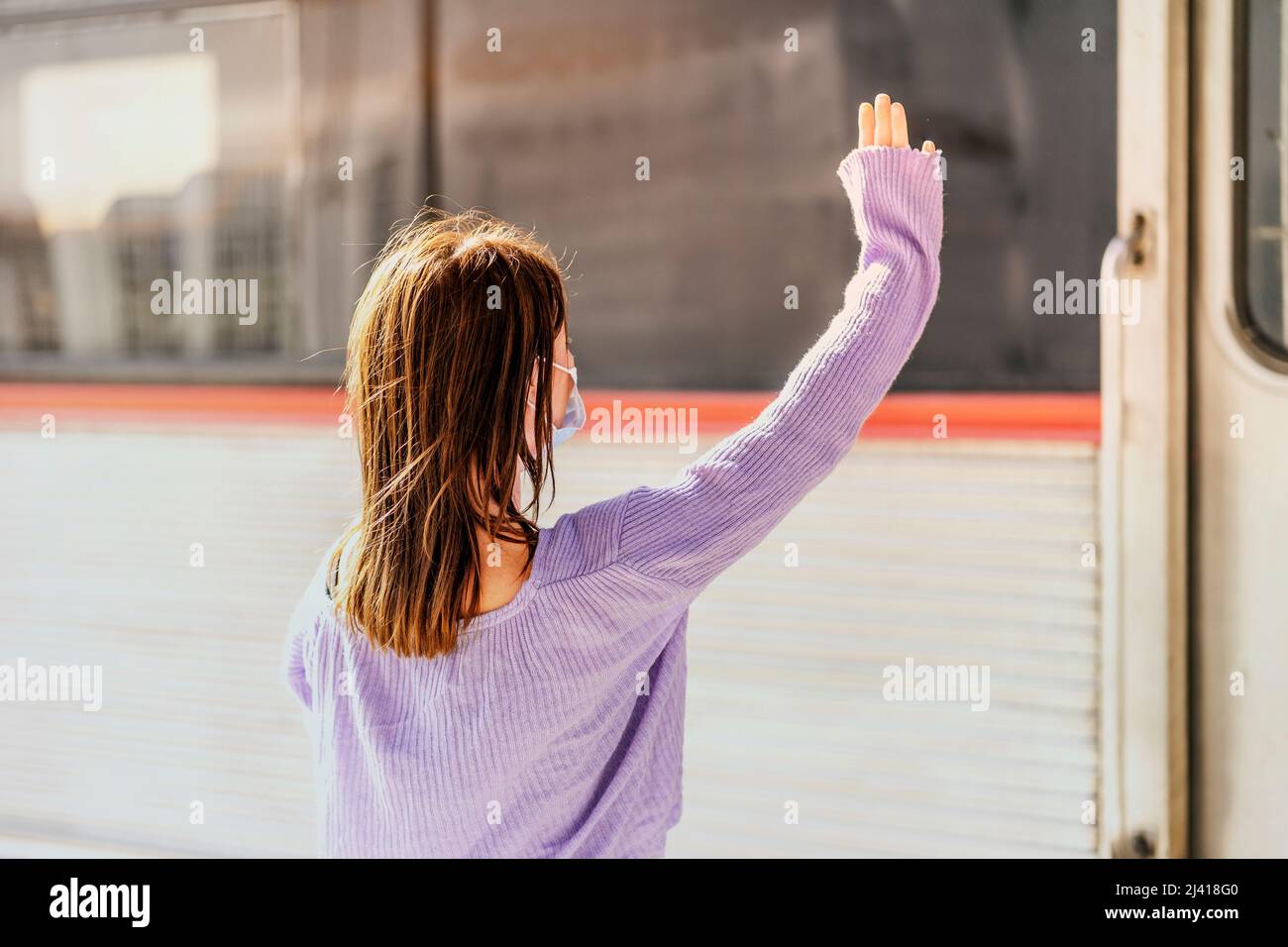 A young woman in a mask waves to somebody in the train from the platform of the railway station Stock Photo