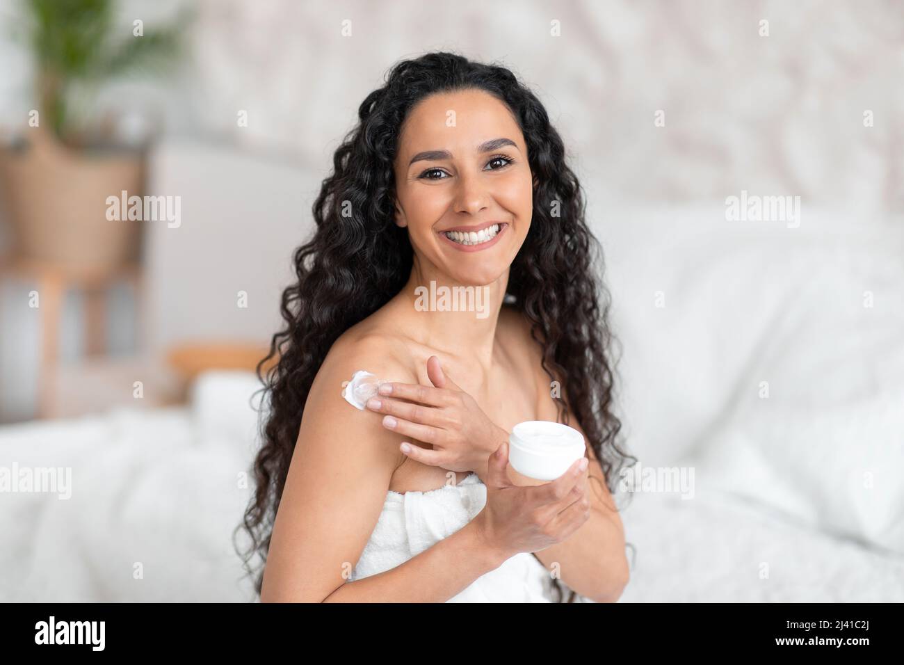 Smiling attractive millennial european brunette woman with long hair in towel applies cream on body in bedroom Stock Photo