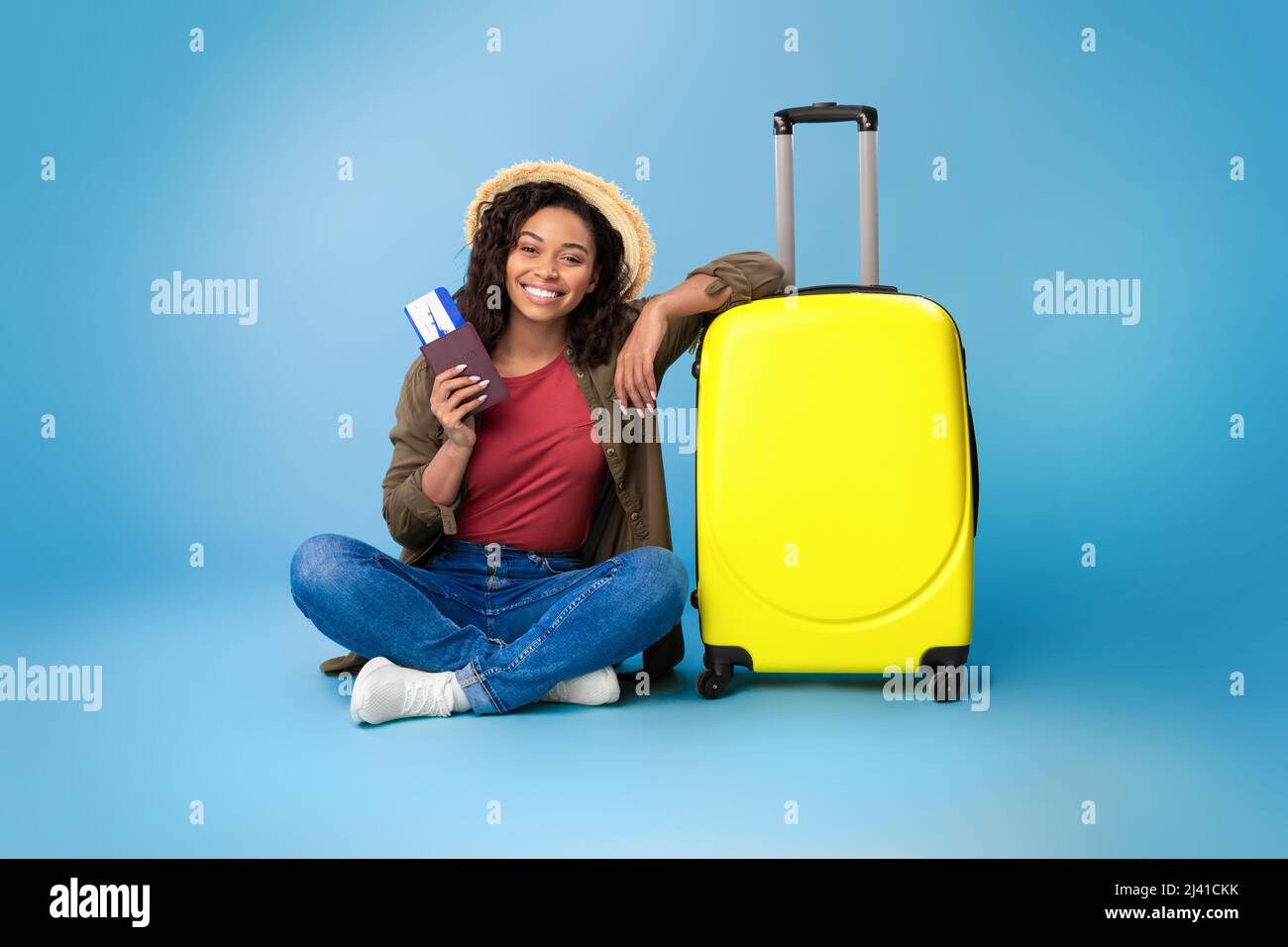 Lovely young black woman holding passport and plane tickets, sitting near bright yellow suitcase on blue background Stock Photo