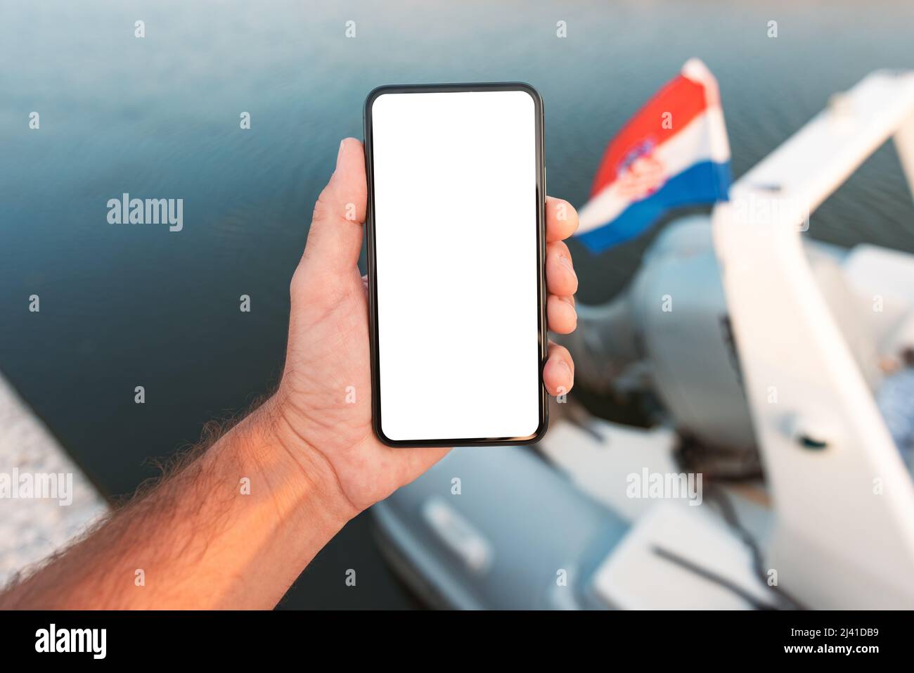 Male hand with mobile smart phone in front of sailing boat with croatian flag, blank mockup screen, selective focus Stock Photo