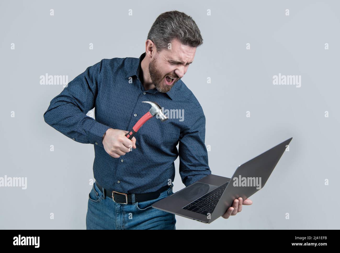 Furious man break laptop hitting notebook with hammer grey background, computer rage Stock Photo