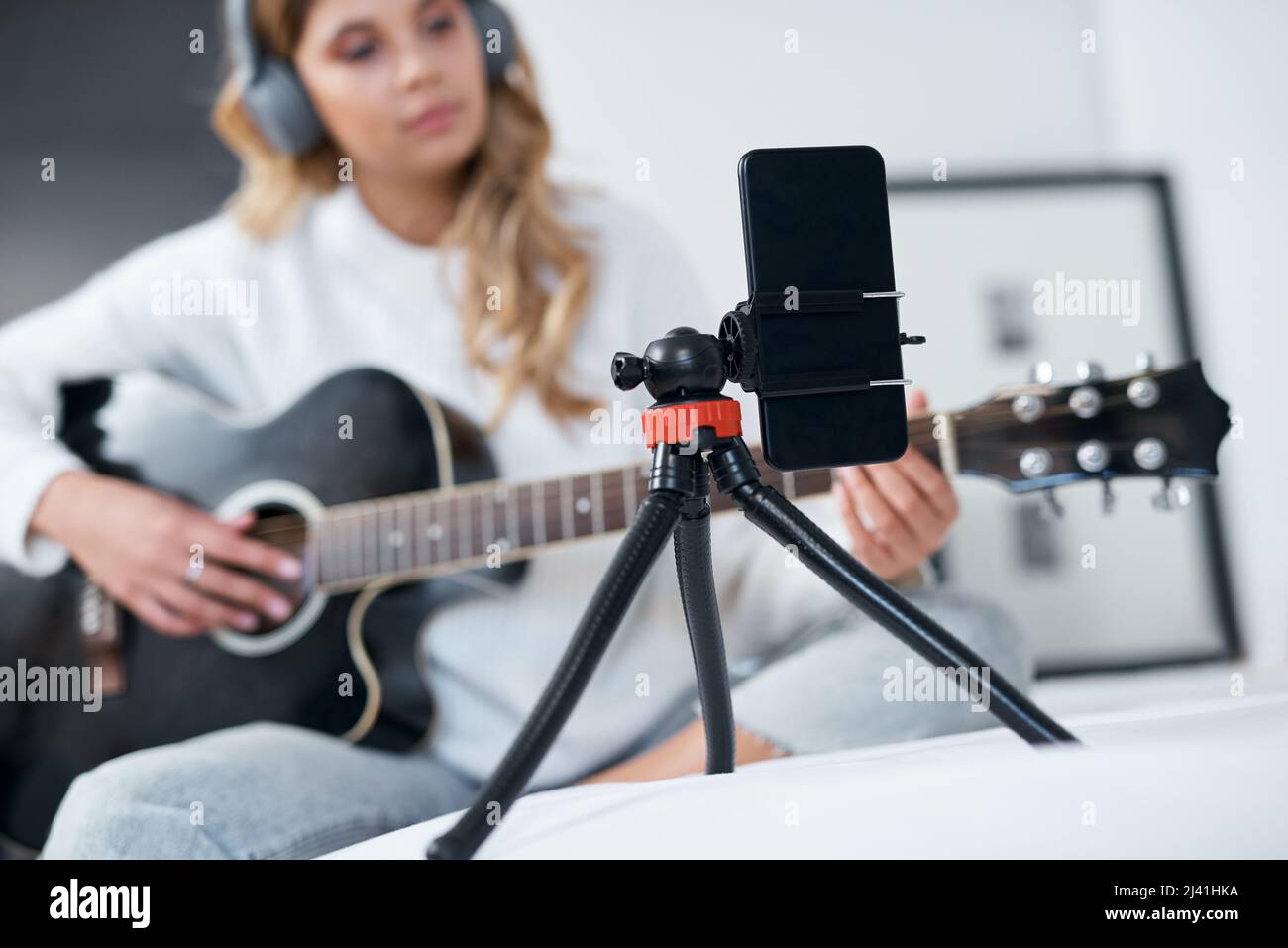 My followers requested this song. Shot of a young woman using her cellphone to record herself while playing the guitar at home. Stock Photo