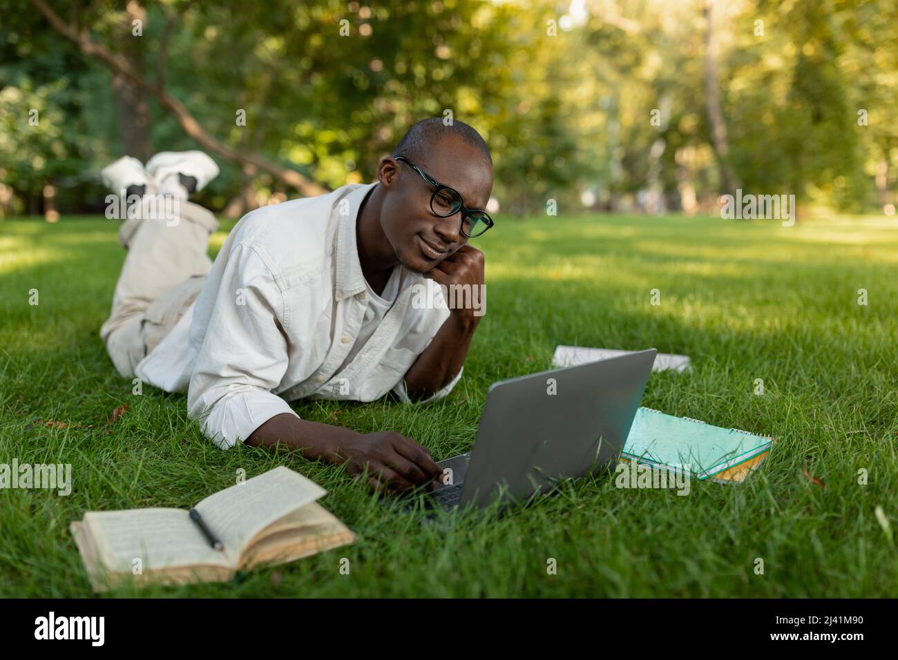 Pensive african american male student getting ready for complicated exam with laptop, lying on lawn in park Stock Photo