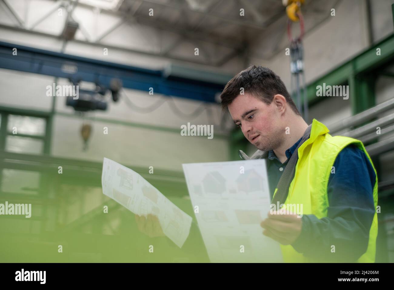 Young man with Down syndrome looking at blueprints when working in industrial factory, social integration concept. Stock Photo