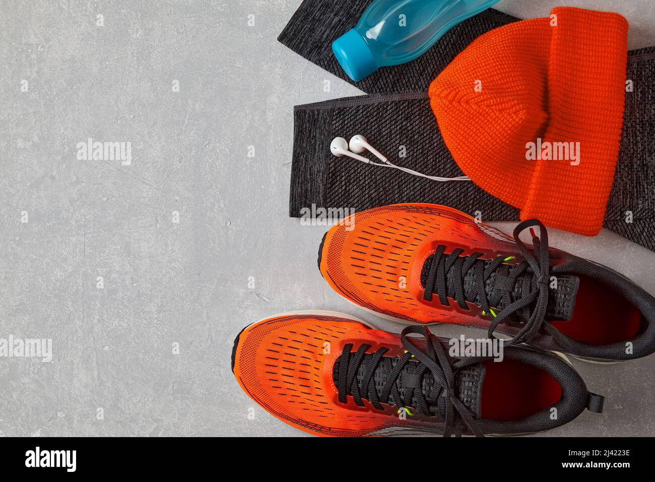 Orange sneakers, an orange knitted hat, warm leggings for sports, a bottle of water and white headphones on a light concrete table. Winter running equ Stock Photo